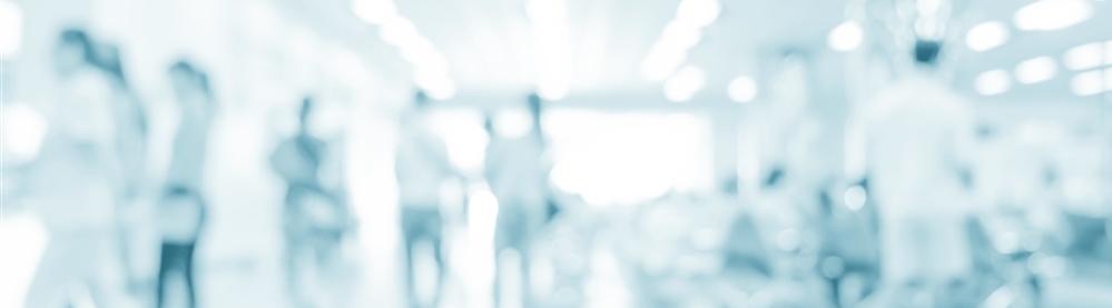 Blurred image of what appears to be people standing and walking around a hospital hallway or waiting room